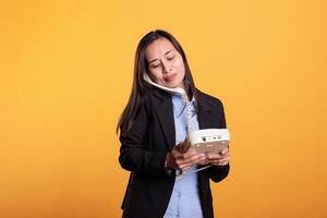 Asian woman in formal suit talking on landline phone call, using telephone with cord standing over yellow background. Confident young adult using office retro phone having remote discussion in studio photo