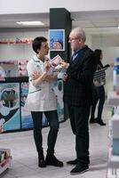 Senior man choosing vitamin for heart health in drugstore and asking pharmaceutical specialist for recommendation. Pharmacist and customer discussing cardiovascular disease prevention in apothecary photo