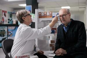 Pharmacist measuring old patient body temperature with thermometer gun, doing health check up and medical consultation. Pharmaceutical specialist testing senior man in glasses for fever in drugstore photo