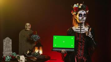 Lady of dead holding laptop with greenscreen, showing isolated copyspace display on pc. Halloween model with creepy body art having isolated chroma key mockup template in studio. photo