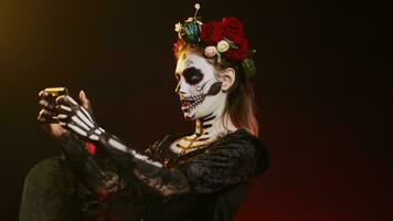 Creepy young woman playing video games on mobile phone, using mobile phone online on dios de los muertos holy tradition. Santa muerte model having fun on scary mexican festival. Handheld shot. photo