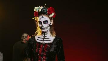 Holy goddess of death posing in halloween costume with skull make up to celebrate dios de los muertos on mexican holiday. Spooky woman as santa muerte with black and white body art. Handheld shot. photo