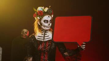 Female model showing isolated speech bubble cardboard, wearing santa muerte costume and skull make up. Person using blank copyspace template on carton board, holy goddess of death. Handheld shot. photo