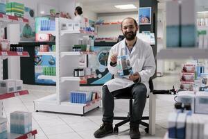 sonriente farmacéutico en pie en silla en farmacia participación médico volantes a promover salud cuidado apoyo servicio. farmacia lleno con farmacéutico productos, vitaminas y drogas foto