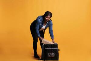 African american employee holding cardboard full with pizza ready to deliver to customer during lunch time in studio with yellow background. Pizzeria courier delivering takeaway food meal photo