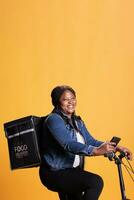 African american courier riding bike while delivering takeaway food meal to client, checking adreess on phone takeout app, standing over yellow background in studio. Take out service and concept photo