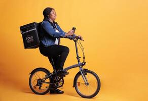 Restaurant employee carrying thermal takeaway backpack while delivering takeout food order to client, holding phone to check address on fast food app. Courier standing over yellow background in studio photo