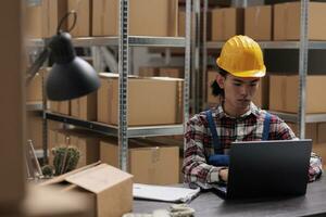 Delivery service worker managing parcel shipment on laptop while working in industrial warehouse. Asian storehouse employee in yellow hardhat analyzing goods supply schedule on computer photo