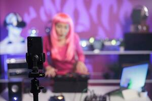 mujer con rosado pelo ejecutando electrónico música utilizando profesional mezclador consola mientras grabación proceso con teléfono inteligente cámara. sonriente artista mezcla canción durante noche hora en club foto