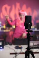 artista con rosado pelo ejecutando electrónico canción a tocadiscos mientras rodaje música proceso con teléfono cámara. DJ mujer haciendo actuación a Club nocturno con profesional audio equipo foto