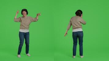 Cheerful young adult dancing and listening to songs on headphones, having fun with mp3 music and dance moves in studio. Female model enjoying sounds on headset, full body greenscreen. photo