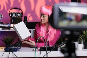 Vlogger with pink hair holding white box while recording product review using professional equipment, sitting at desk in filming studio. Content creator talking with subscribers during live streaming photo