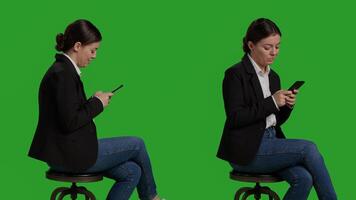 Close up of businesswoman browsing internet on mobile phone, acting casual on chair in studio. Company worker in suit using smartphone app to text messages, full body greenscreen backdrop. photo