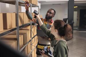 Storehouse supervisor showing colleague invoice code on device to find clients parcel in storage room. Shipping service employees scanning merchandise products while analyzing logistics report photo