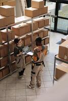 Warehouse supervisor working at products inventory on tablet computer, inspecting goods during quality control in storage room. Shipping company workers packing boxes, discussing cargo audit. Top view photo