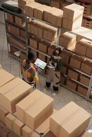 Diverse storehouse team scanning cardboard boxes, checking online products inventory on digital tablet. Storage room employees in protective overalls preparing orders, working in storehouse. Top view photo