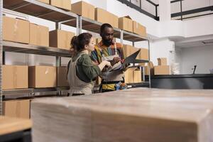 Storage room coworkers searching parcel using checklist on digital tablet, preparing cardboard boxes for shipping. Entrepreneur wearing overalls scanning cardboard box using store scanner photo