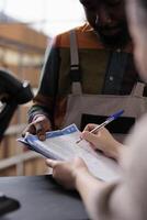 Supervisor signing merchandise quality control report, discussing customers online orders before start preparing packages. Diverse team analyzing goods checklist in warehouse. Close up photo