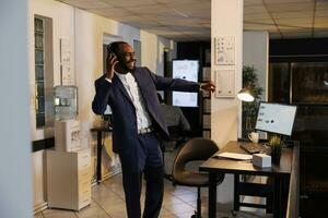 Smiling entrepreneur listening music during work break, dancing in startup office after finishing company strategy. African american businessman enjoying having fun late at night in startup office photo