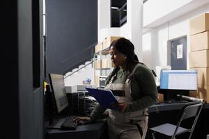 Stockroom manager working at merchandise inventory, analyzing products checklist on computer in storehouse. African american employee preparing customers orders during goods inventory photo