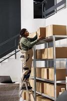 Stockroom worker standing on ladder putting cardboard box on shelf, preparing customers orders in storage room. African american storage room supervisor working at merchandise quality control photo