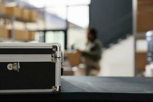 Selective focus of metallic box standing on counter desk in warehouse, in background african american worker preparing customers packages. Stockroom manager working at merchandise inventory photo