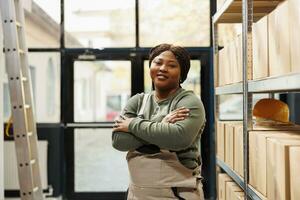 Warehouse worker standing with arm crossed smiling at camera while working at goods inventory. African american employee preparing customers order before strat delivery packages in storehouse photo