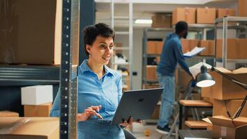 Warehouse owner checking cardboard boxes from storage space shelves, using laptop to plan stock inventory and take notes. Entrepreneur working on startup development and supply chain. photo