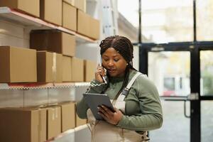 Supervisor answering landline phone call in storage room, talking with remote manager about products quality control. African american employee with overall while checking logistics on device photo