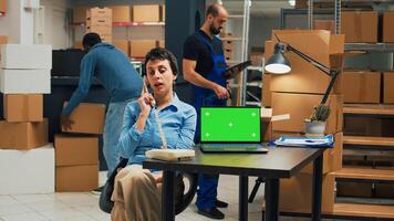 Young woman answering landline phone call in storage room and using greenscreen display on laptop. Person talking on telephone with cord and looking at blank mockup copyspace. photo