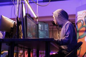 Internet criminal in hood planning online scam while coding malicious software on computer. Asian hacker using malware to breach cybersecurity system and steal privacy data photo