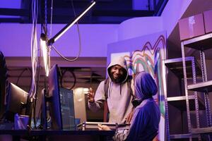 Men in hood discussing hacking plan with cheerful smile in abandoned warehouse. Happy young african american hacker explaining illegal ransomware code on computer to developer photo