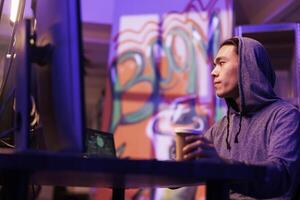 Hacker in hood cracking database password and drinking takeaway coffee. Young asian criminal looking at computer screen and holding beverage to go while coding internet virus malware photo