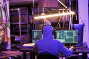 Hacker in headphones listening to music while cracking password on computer. Internet criminal in earphones planning online fraud and coding spyware in hideout place with neon light photo