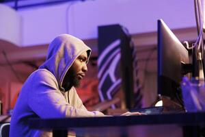 Scammer using malicious software to break into database and steal valuable information. African american hacker in hood planning phishing fraud on computer in hideout at night photo