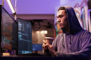 Asian hacker drinking coffee and doing ddos attack on website. Criminal breaking into network system on computer screen while holding beverage to go in abandoned warehouse photo