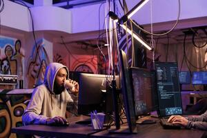 African american hacker drinking coffee and doing illegal activities late. Criminal holding takeaway cup while hacking server and coding malware in abandoned warehouse at night photo