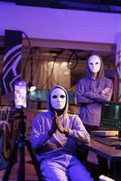 Hackers in masks recording threatening message and demanding victim for confidential privacy information. Anonymous internet scammers doing criminal activities and streaming on mobile phone camera photo