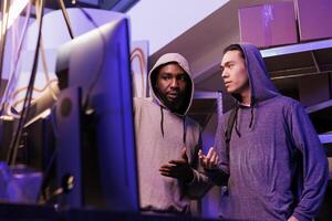 Hackers discussing malware code while looking at computer screen. Two young men in hoods planning illegal activity and talking about network system breaking at night time photo