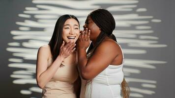 African american woman whispering secret to asian girl on camera, acting sensual and glamorous in studio. Interracial skincare models promoting body positivity and acceptance, having fun. photo