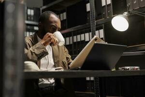 African american cop drinking coffee and reading csi report at night time. Detective sitting at workplace desk, holding tea mug and studying crime case file in dark office room photo