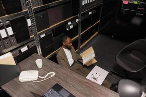 Overworked exhausted cop sitting on floor analyzing report in office. Stressed african american policeman reading crime case materials papers and working late at night time top view photo