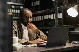 Worried detective analyzing crime case on laptop and thinking about evidence. Concerned african american policeman sitting at workplace desk in office full of racks with folders and files photo