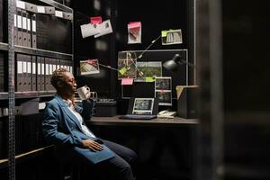 Pensive african american woman investigator drinking coffee, analyzing murder case. Thoughtful woman holding tea cup, sitting at detective desk while studying clues at night time photo