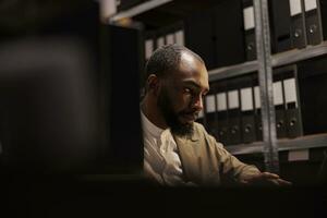 African american cop conducting investigation in police archive, sitting near shelf full of folder with records. Concentrated private detective solving complex crime case photo