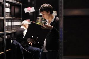 Fbi woman agent holding confidential crime files, working overtime at victim report in arhive room. Criminology workers analyzing suspect information, discussing criminal investigations strategy photo
