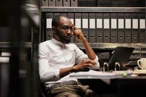 African american detective solving complex crime case using laptop. Law enforcement professional thinking about evidence and checking proofs on computer in police office room photo