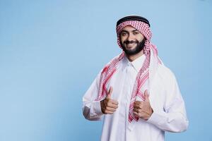 Arab man wearing islamic clothes giving thumbs up and showcasing cheerful expression portrait. Smiling muslim person dressed in traditional robe and headscarf and looking at camera photo