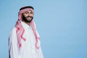 Cheerful muslim man with carefree expression dressed in traditional arabian clothes looking at camera. Smiling person wearing checkered red and white headdress studio portrait photo