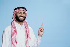 Cheerful muslim man dressed in traditional robe pointing upwards with smiling expression. Carefree arab advertising product while showing in up direction and looking at camera photo